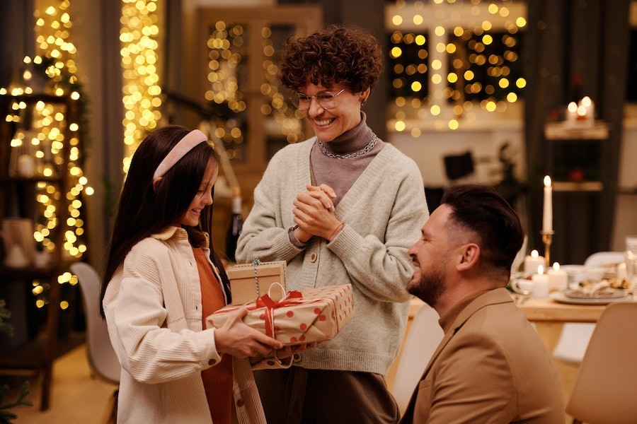 A man and a woman smile while their daughter happily shows them her Christmas presents, still wrapped.